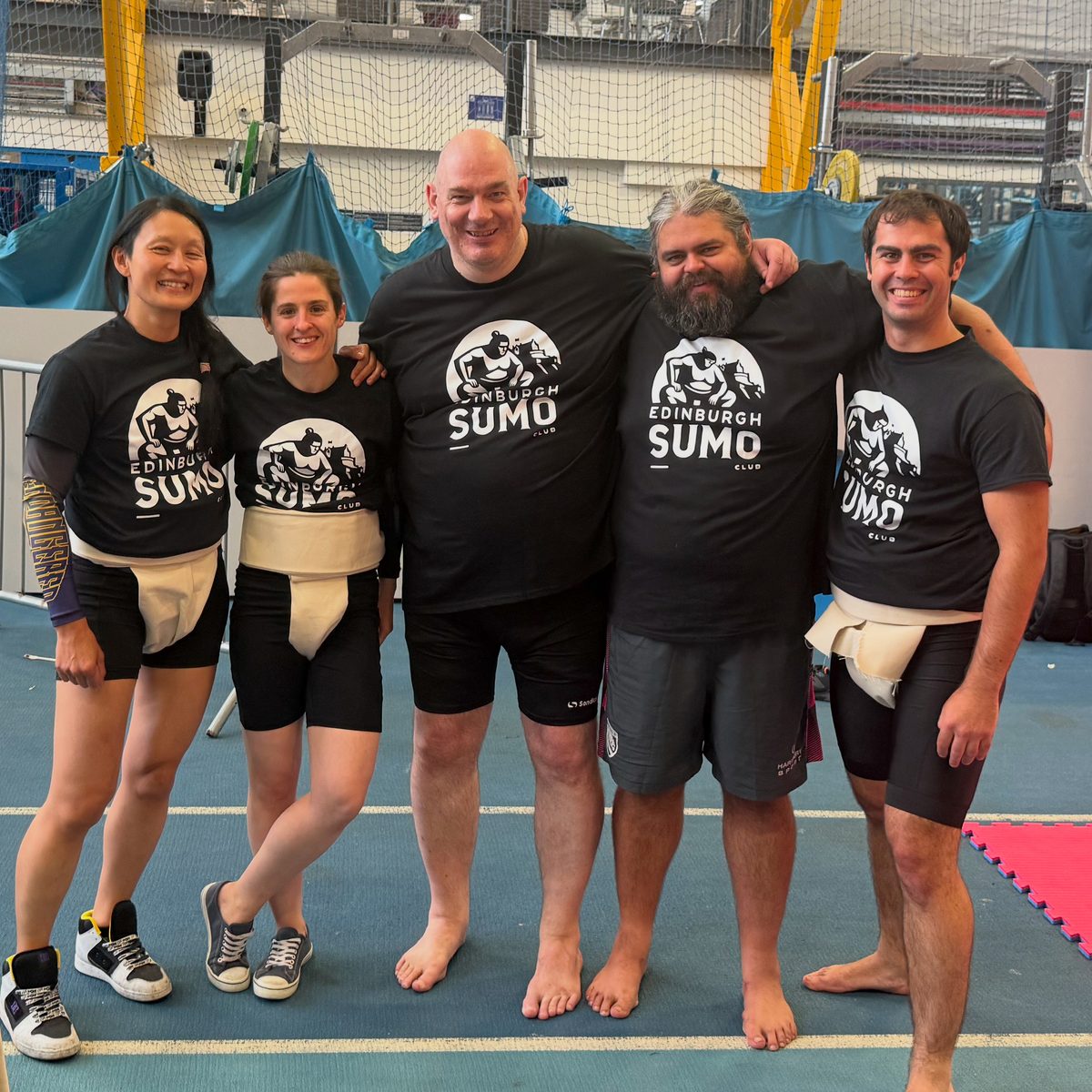 Five club members in mawashi and club t-shirts at a competition venue.